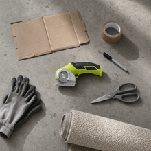 Overhead flatlay on a garage floor: the cutter with a flattened box, tape, marker, gloves and the blunt scissors it replaces
