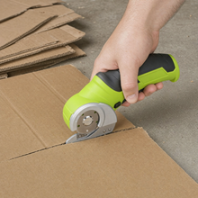 The cutter going through a thick flattened moving box on a garage floor, beside a stack of already flattened boxes